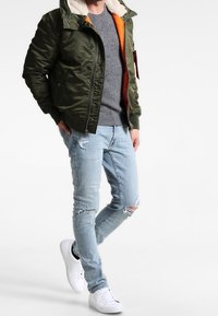 Blouson aviateur vert olive avec col en peau lainée, doublure orange, pull gris, jeans bleu clair déchirés et baskets blanches.