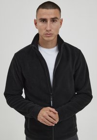 Chaqueta de forro polar negra con cremallera y cuello alto. Presenta mangas largas y una textura suave, exhibida sobre un fondo neutro.