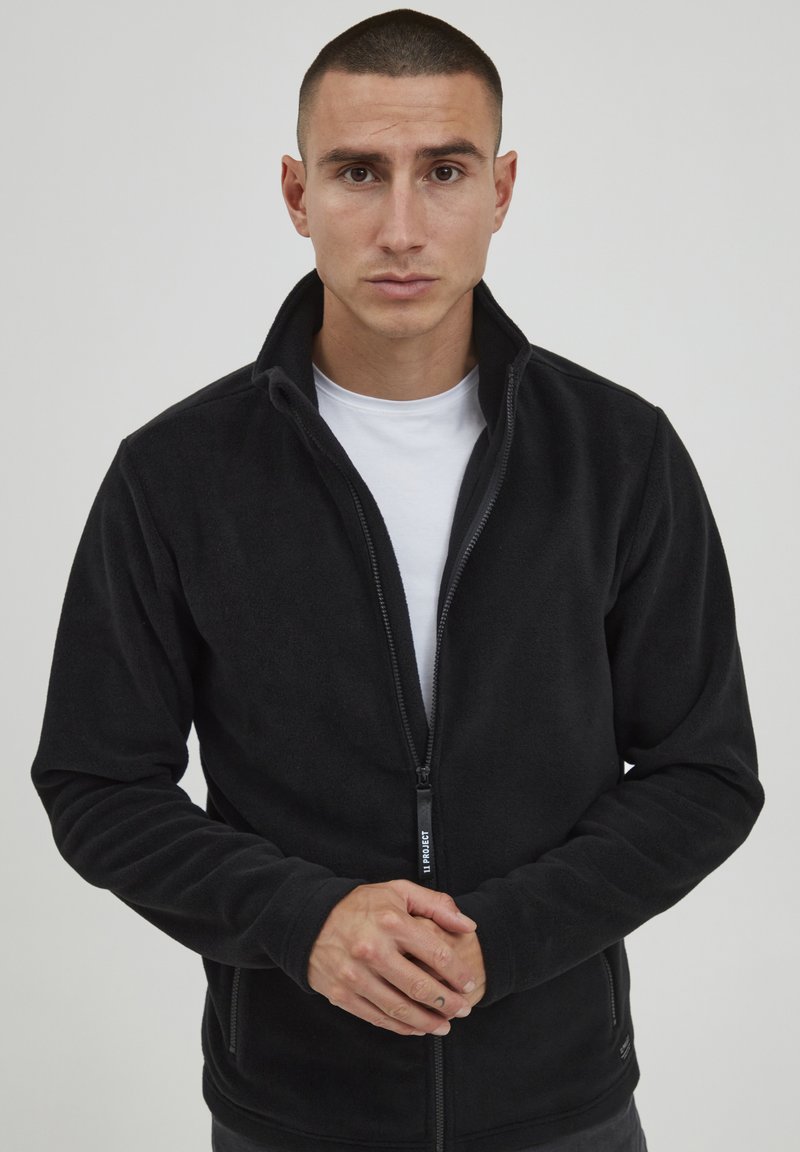 Svart fleece zip-up jakke med høy krage og glidelås. Har lange ermer og en myk tekstur, vist på en nøytral bakgrunn.