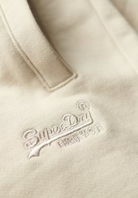 Tessuto di polo beige con una finitura texturizzata, caratterizzato dal logo ricamato "SuperDry" in evidenza vicino al colletto.