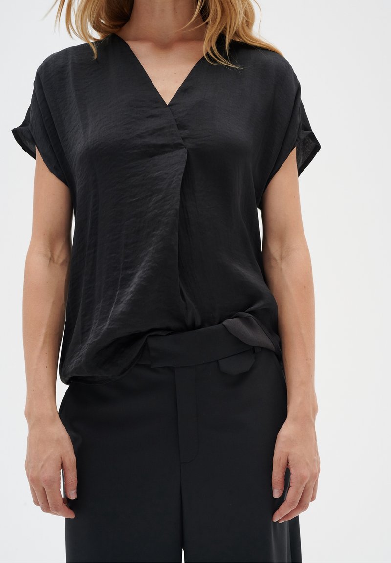 Haut noir à manches courtes avec un col en V et une texture froissée, présentant une coupe ample et un design drapé, associé à un pantalon noir ajusté.