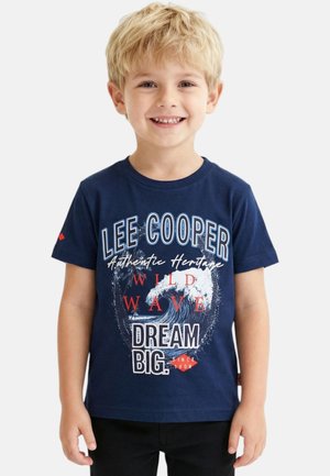 Jeune garçon blond souriant, portant un t-shirt bleu marine avec un texte blanc et rouge ainsi qu'un graphisme de vague, debout devant un arrière-plan clair uni.