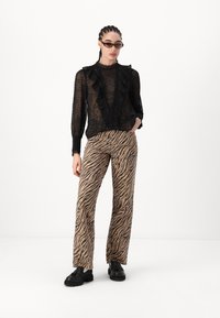 Zwarte doorzichtige blouse met ruches bij de hals, gecombineerd met lichtbruine broeken met zwarte tijgerstrepen. Gestyled met zwarte plateauschoenen.