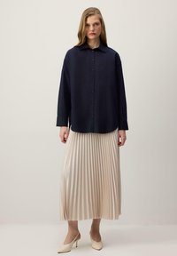 Femme portant une chemise bleu marine à boutons, une jupe plissée midi beige et des escarpins slingback crème, debout devant un fond clair uni.