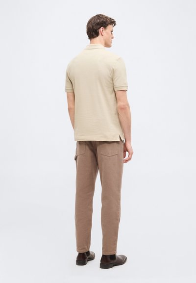 Homme portant un polo à manches courtes beige, un pantalon marron avec poche cargo, et des chaussures marron foncé, debout, de dos sur un fond clair uni.