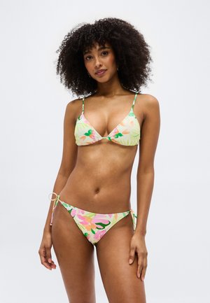 Žena s kučeravými vlasmi nosiaca pastelové kvetinové bikiny s tenkými ramienkami, stojaca pred jednoduchým svetlým pozadím.