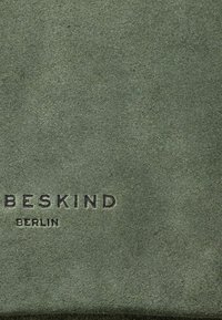 Materiale in suede verde con una texture morbida, caratterizzato da testo in rilievo "BESKIND BERLIN". L'aspetto complessivo è minimalista e pulito.