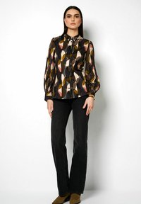 Mehrfarbige Bluse mit geometrischem Muster in Braun, Gelb und Pink, mit Knopfleiste und langen Ärmeln. Kombiniert mit schwarzen Jeans.