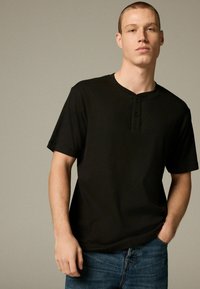 Sort kortærmet Henley t-shirt med tekstureret finish, der har tre knapper ved halsudskæringen og en afslappet pasform, parret med blå jeans.