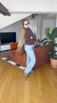 Ung kvinne med solbriller og skjerf poserer ved en brun sofa i et lyst stue med tregulv og stor potteplante.