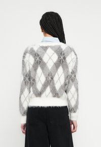 Műszőrme pulóver szürke és fehér gyémántmintával, rövidített fazon, puha tapintású, bordázott mandzsettákkal. Fekete nadrággal párosítva.