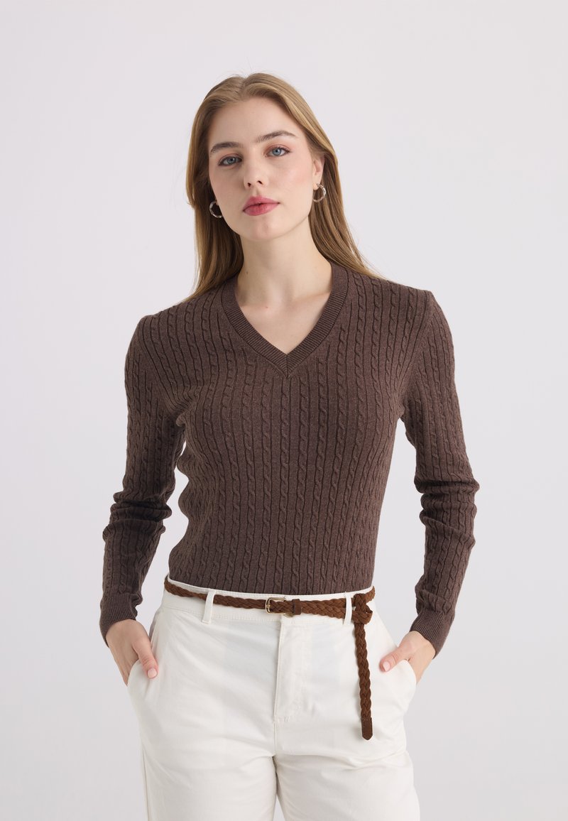 Femme aux longs cheveux blonds portant un pull marron en maille torsadée à col en V, un pantalon blanc et une ceinture marron tressée, les mains dans les poches.