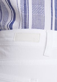 Jeans blancos con una textura suave y un parche de cuero rectangular que presenta "OPUS". Tela a rayas azul y blanca parcialmente visible arriba.