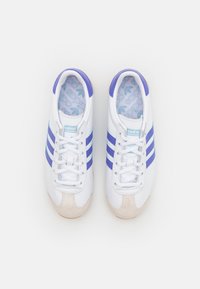 Bílé sportovní boty s purpurovými pruhy a akcenty, s texturovanou špičkou, zaobleným designem a kontrastní světle modrou stélkou.
