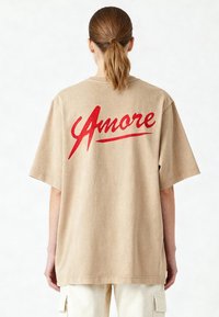 Bež majica s kratkimi rokavi v oversize stilu, okroglo izrezanega vratu, ki na hrbtni strani izstopa s poudarjenim rdečim napisom "Amore".