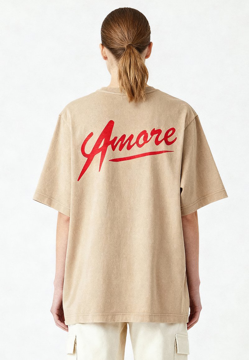 Bež majica s kratkimi rokavi v oversize stilu, okroglo izrezanega vratu, ki na hrbtni strani izstopa s poudarjenim rdečim napisom "Amore".