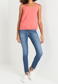 Korálový top bez rukávů s kulatým výstřihem, kombinovaný s světle modrými skinny džíny a bílými nazouvacími botami. Top má detail na shromážděném pase.
