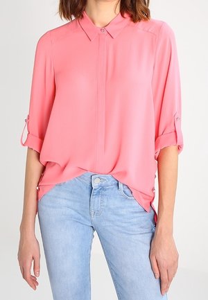 Pink, lightweight blouse with a collar, roll-up sleeves, and a loose fit. Paired with light blue denim jeans.