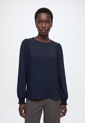Femme aux cheveux courts portant un chemisier bleu marine texturé à manches longues et un pantalon gris, debout devant un fond blanc uni.