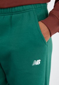 Žalios medvilninės sportinės kelnės su elastinga juosmeniu, šoninėmis kišenėmis ir balta New Balance logotipu kairėje apatinėje kojoje.