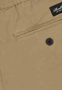 Tejido beis con una textura suave que presenta un bolsillo horizontal con cierre de botón y una etiqueta negra cosida en la cinturilla.