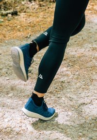 Scarpe da ginnastica blu con tomaia testurizzata e suola bianca, abbinate a leggings neri, su un sentiero in ghiaia con foglie sparse.