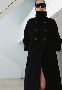Czarny, dwurzędowy wełniany płaszcz z wysokim kołnierzem, z dużymi guzikami i bocznym rozcięciem. Gładka faktura i dopasowany krój.