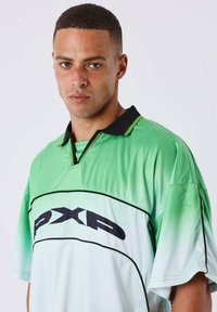 Giovane uomo con capelli corti che indossa una maglia sportiva PXP verde e bianca con colletto nero, che guarda direttamente verso la fotocamera.