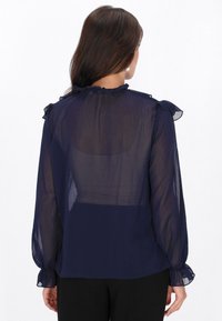 Tengerészkék áttetsző blúz fodros díszítéssel a vállakon és az ujja szegélyén, hosszú ujjal és magas nyakkal. Simább, könnyű anyag.