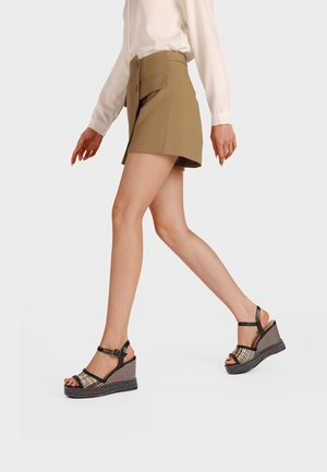 Donna che cammina con gonna color camoscio e camicetta bianca, indossando sandali con zeppa neri e beige con cinturini alla caviglia su uno sfondo semplice.