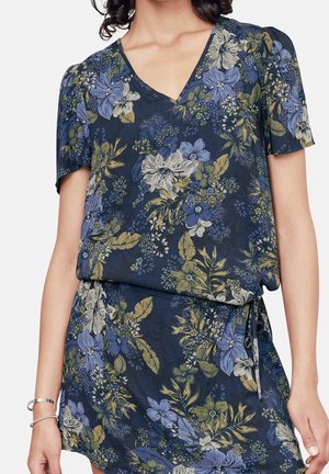 Vrouw draagt een marineblauwe jurk met korte pofmouwen en een V-hals, met groene, witte en paarse bloemenpatronen.