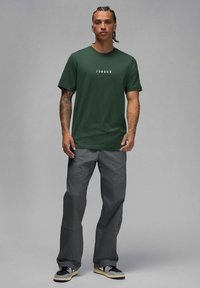 Camiseta de algodón verde con el texto blanco "JORDAN", combinada con pantalones grises de corte holgado y costuras distintivas. Zapatillas negras y grises.