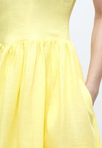 Κίτρινο φόρεμα χωρίς μανίκια με σφηκοειδή μέση, υφασμάτινη υφή και χέρι τοποθετημένο μέσα στην πλαϊνή τσέπη σε φωτεινό φόντο.