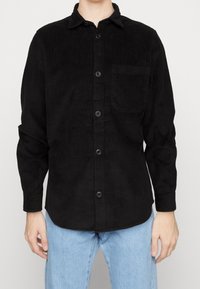 Musta corduroy-paita, jossa on napillinen etuosa, terävä kaulus, pitkät hihat ja rintatasku, jossa on näkyvät pystysuuntaiset uritukset.