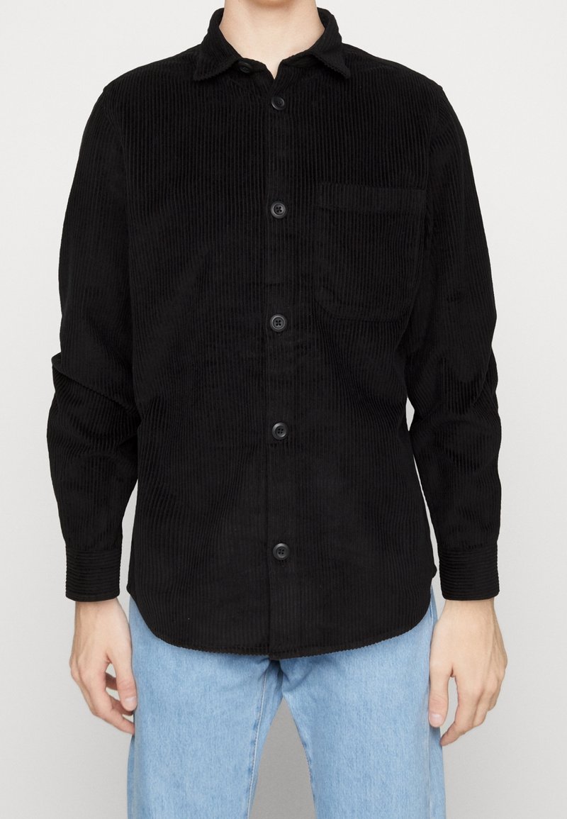 Musta corduroy-paita, jossa on napillinen etuosa, terävä kaulus, pitkät hihat ja rintatasku, jossa on näkyvät pystysuuntaiset uritukset.