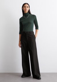 Grüner Rollkragenpullover aus weichem Stoff, kombiniert mit schwarzen weit geschnittenen Hosen mit Nadelstreifen. Das Model steht mit den Händen in den Taschen und hat nur minimale Accessoires.