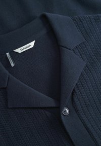 Mörkblå stickad polo skjorta med ett texturerat mönster, platt krage och en enda knapp. Etiketten visar "GABBA" på en vit tag.