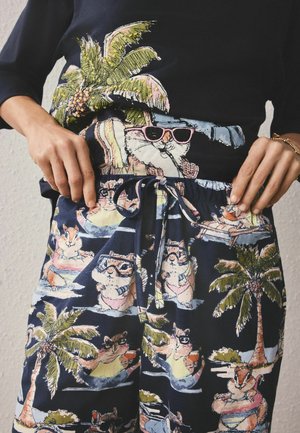 Person iført navy outfit med jordegern i solbriller, der slapper af på poolflåder med palmer, holder i snørrebåndet på taljen.