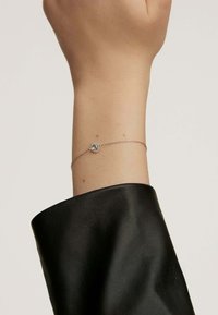 Pulsera de cadena de plata con un centro de piedra clara y redonda, llevada en una muñeca parcialmente cubierta por una manga de cuero negro.