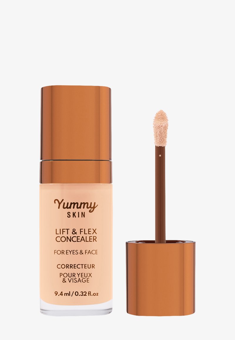 Correcteur liquide beige dans un flacon transparent avec un capuchon en bronze. Équipé d'un applicateur en forme d'œuf, étiqueté "Yummy Skin Lift & Flex Concealer."