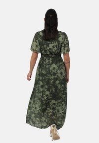 Maxi-Kleid in Dunkelgrün mit floralem Print. Durchsichtiger Stoff, kurze Puffärmel, elastischer Taillenbund, fließende Silhouette, getragen mit hellen Schuhen.
