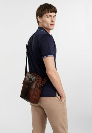 Mężczyzna w granatowym polo i beżowych spodniach, noszący brązową skórzaną torbę na ramię, stojący na białym tle.
