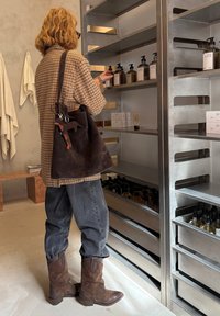Una persona che seleziona prodotti da un'unità di scaffali in metallo. Indossa una camicia a quadri marrone, jeans scuri e stivali da cowboy marroni. Una borsa scura pende dalla sua spalla.