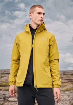 Hombre con una chaqueta con capucha amarilla mostaza sobre una camisa negra con cremallera, de pie al aire libre sobre un terreno rocoso bajo un cielo nublado.