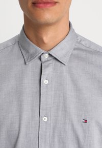 Camisa gris abotonada con tela texturizada, que presenta un cuello, botones blancos y un pequeño logo en rojo, blanco y azul en el lado izquierdo del pecho.