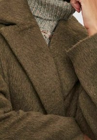 Vlněný kabát olivově zelené barvy s texturovaným povrchem, širokým klopovým límcem a volným střihem. Svetr pod ním je žebrovaný a vícebarevný.