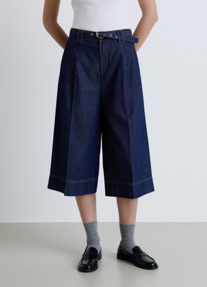 Persona che indossa pantaloni larghi blu scuro corti con cintura, calzini grigi e mocassini neri, in piedi davanti a uno sfondo chiaro e uniforme.