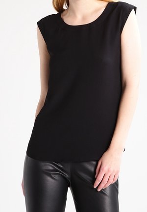 Kobieta ubrana w czarną, bezrękawową bluzkę i czarne skórzane spodnie, stojąca z jedną ręką opartą na udzie na białym tle.