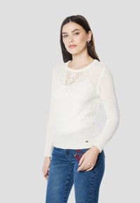 Roomkleurige gebreide trui met een gestructureerd patroon, voorzien van een kanten inzet bij de halslijn. Lange mouwen en een aansluitend ontwerp. Gecombineerd met blauw geborduurde spijkerbroek.