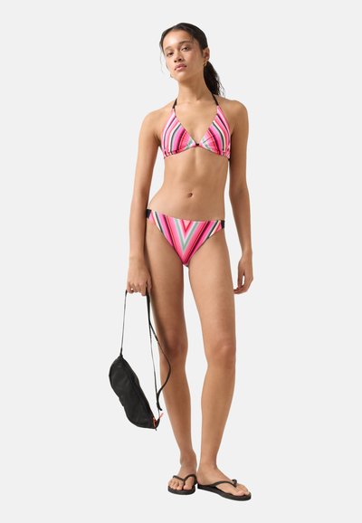 Jeune femme en bikini rose à rayures tenant un sac noir, portant des tongs noires, debout devant un fond clair uni.